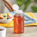 A 11.5 oz. Skep PET sauce/honey bottle with a white flip top lid on a table with a bowl of fruit.