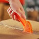 A hand using a CrewSafe orange plastic utility knife with an orange blade to cut a piece of plastic.
