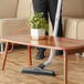 A person using a Lavex backpack vacuum to clean a coffee table with a plant on it.