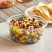 A Stalk Market clear deli container filled with corn and black bean salsa with tortilla chips on a table.