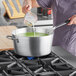 A woman pouring green liquid into a Vollrath Wear-Ever sauce pan on a stove.