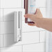 A person using a white DoveLok bottle bracket to hold a bottle of soap on a white tile wall.