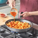 A person pouring red sauce into a Vollrath stainless steel non-stick fry pan of pasta.
