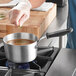 A person pouring soup from a bowl into a Vollrath Wear-Ever sauce pan on a gas stove.
