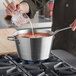 A hand pouring milk into a Vollrath stainless steel sauce pan on a stove.