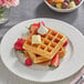 A plate with Krusteaz Belgian waffles, butter, and strawberries with a bowl of fruit.