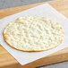 A round white CAULIPOWER pizza crust on white paper on a wooden cutting board.