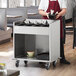 A ServIt stainless steel tray cart with utensils and bowls on wheels in a professional kitchen.