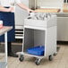 A person in a blue apron using a ServIt stainless steel flatware tray cart to hold a tray of flatware containers.