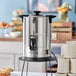An Avantco stainless steel coffee urn on a table with a cup of coffee.