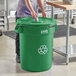 A woman in a kitchen putting a Lavex green recycling can with a bottle lid into a green trash can.
