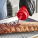 A hand using a Vollrath red bottle cap to pour barbecue sauce on a rack of ribs.