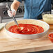 A person using a spoon to spread red sauce on a pizza in a Choice aluminum deep dish pizza pan.