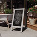 A Flash Furniture Canterbury vintage graywashed wood A-frame chalkboard sign on a sidewalk with text on it.