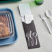 A black paper cutlery caddy with a fork and spoon next to a container of beans and chicken.