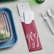 A Dinex burgundy paper cutlery caddy with plastic forks and spoons next to a plastic container.