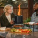 A man and woman sitting at a table with food and a Solo Stove Mesa XL 7" Black Stainless Steel Tabletop Fire Pit with a fire inside.