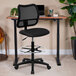 A Flash Furniture black mesh mid-back drafting chair with adjustable foot ring at a desk with a plant.