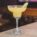 An Arcoroc Romeo margarita glass on a table with yellow liquid and a lime wedge.