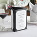 A black Acopa Prime 3-view triangular table tent on a table with wine glasses and a menu.