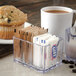 A clear Carlisle plastic sugar caddy on a counter with sugar packets and a cup of coffee.