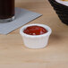 A small white Hall China flared ramekin filled with red sauce on a table next to a glass of soda.