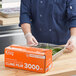 A man in a chef's uniform uses a serrated cutter to dispense Choice Heavy-Duty Foodservice Film into a container.