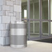 A silver stainless steel cylindrical trash can with holes in it next to a stone wall.