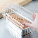 A hand holding a clear plastic dome lid over a foil bread loaf pan of food.