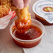 A hand holding a fried chicken stick over a container of Sweet Baby Ray's Sweet Red Chili Wing Sauce.