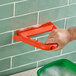 A person using a Carlisle orange grout brush with a red handle to clean a tile wall.