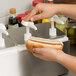 A person using a Carlisle condiment dispenser to put ketchup on a hot dog.