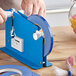 A person using a blue Lavex poly bag sealer tape dispenser to cut tape on a table.