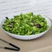 A white GET Siciliano bowl filled with green salad on a table with tongs.