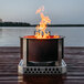 A BREEO stainless steel fire pit base with a fire in it on a wooden deck overlooking water.