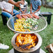 A BREEO stainless steel grill grate with meat and vegetables cooking on it over a fire pit.