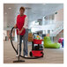 A woman in a red apron using a red and black NaceCare cordless canister vacuum to clean the floor.