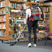 A woman wearing a white shirt and a grey and orange NaceCare cordless backpack vacuum cleaning a book shelf.