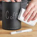 A hand using a white Planetary Design waterproof writer pen to write on a black mug.