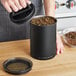 A person pouring dried herbs into a Planetary Design matte black stainless steel food storage container.