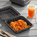 A black plastic hinged take-out container with food in it next to a drink and fork.