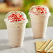 Two Choice clear plastic cups of milkshakes with whipped cream and red sprinkles with dome lids.