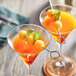 Two glasses of orange liquid with fruit on stainless steel skewers.