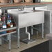 A Regency underbar sink with faucet and 2 drainboards on a counter with bottles above.