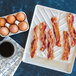 Nordic Ware high-heat plastic bacon tray with bacon, eggs, and coffee on a counter.