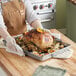 A person wearing gloves and an apron holding a turkey with orange slices on top in a Nordic Ware aluminum roasting pan.