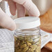 A person in gloves holding a white 53/400 polypropylene cap over a jar of green seeds.