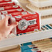 A hand holding a red and white package of Om Sweet Home Plant-Based Vegan Lightly Salted Butter sticks on a refrigerator shelf.