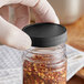 A hand holding a 53/400 black plastic cap on a jar of red pepper flakes.