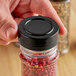 A hand holding a jar of red peppercorns with a black unlined polypropylene spice cap on it.